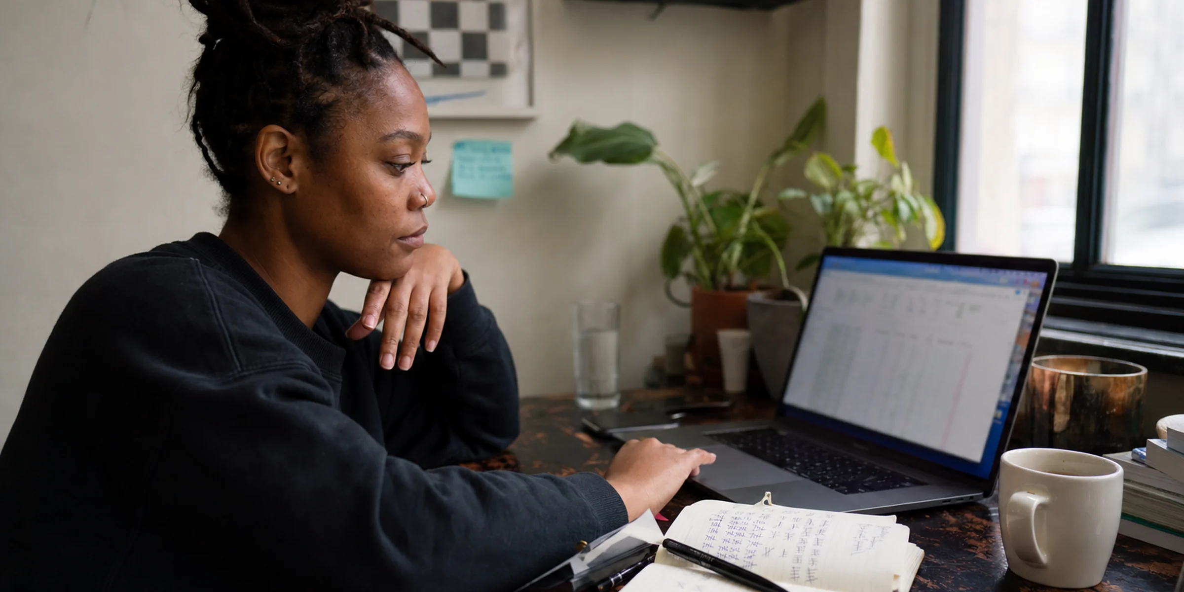 Black woman founder at a home desk reviewing blurred customer metrics on a laptop with cancellation tallies in an open notebook