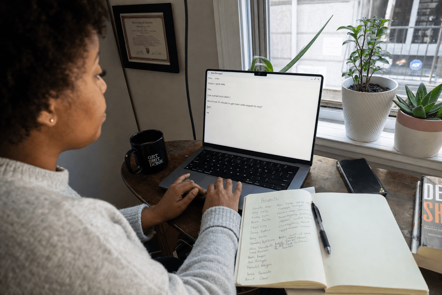 Solo founder at a home desk drafting a short plain-text email on a laptop with a handwritten prospect list in a notebook