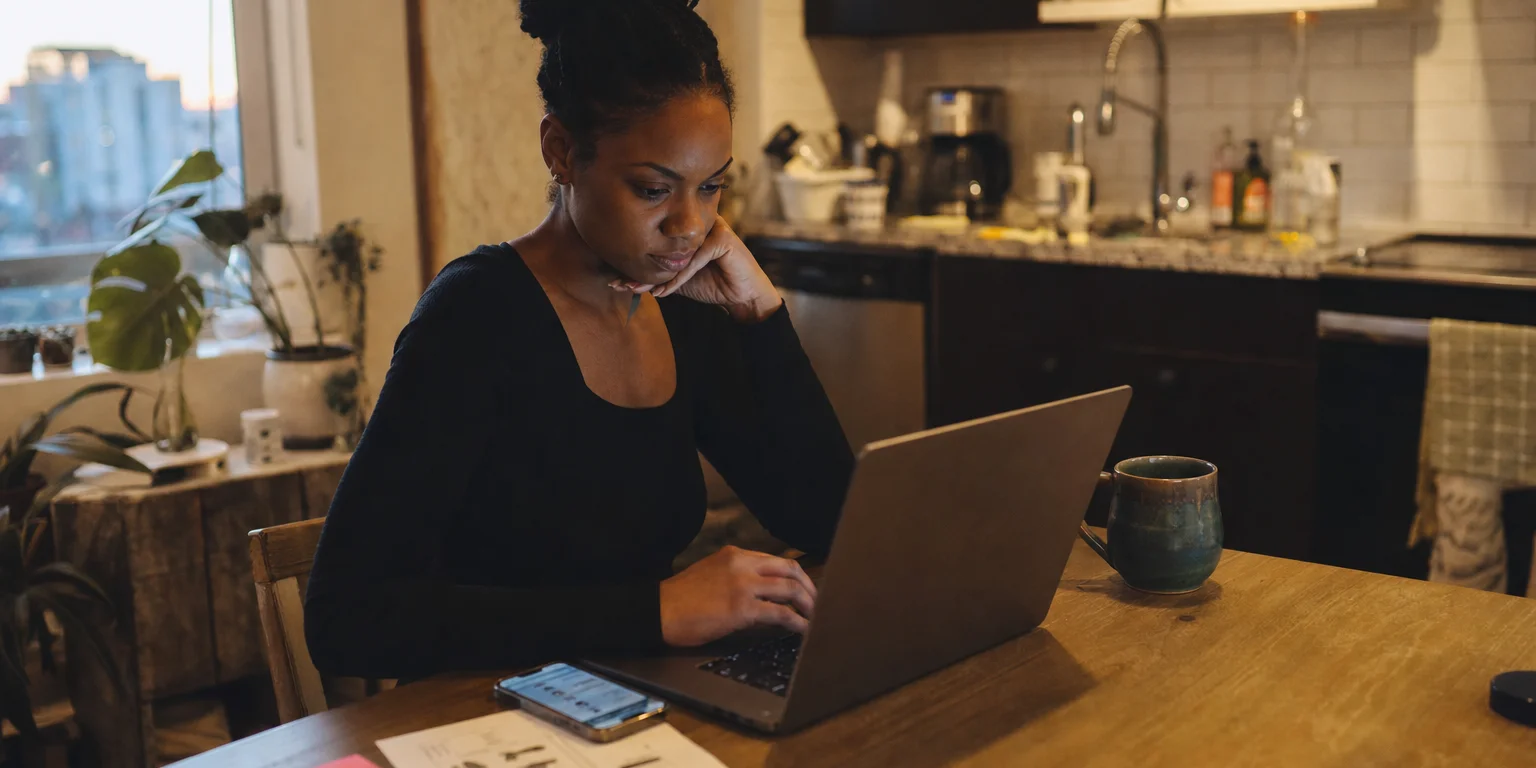 Solo founder at a kitchen table with a laptop, phone, and printed tool checklist beside sticky notes