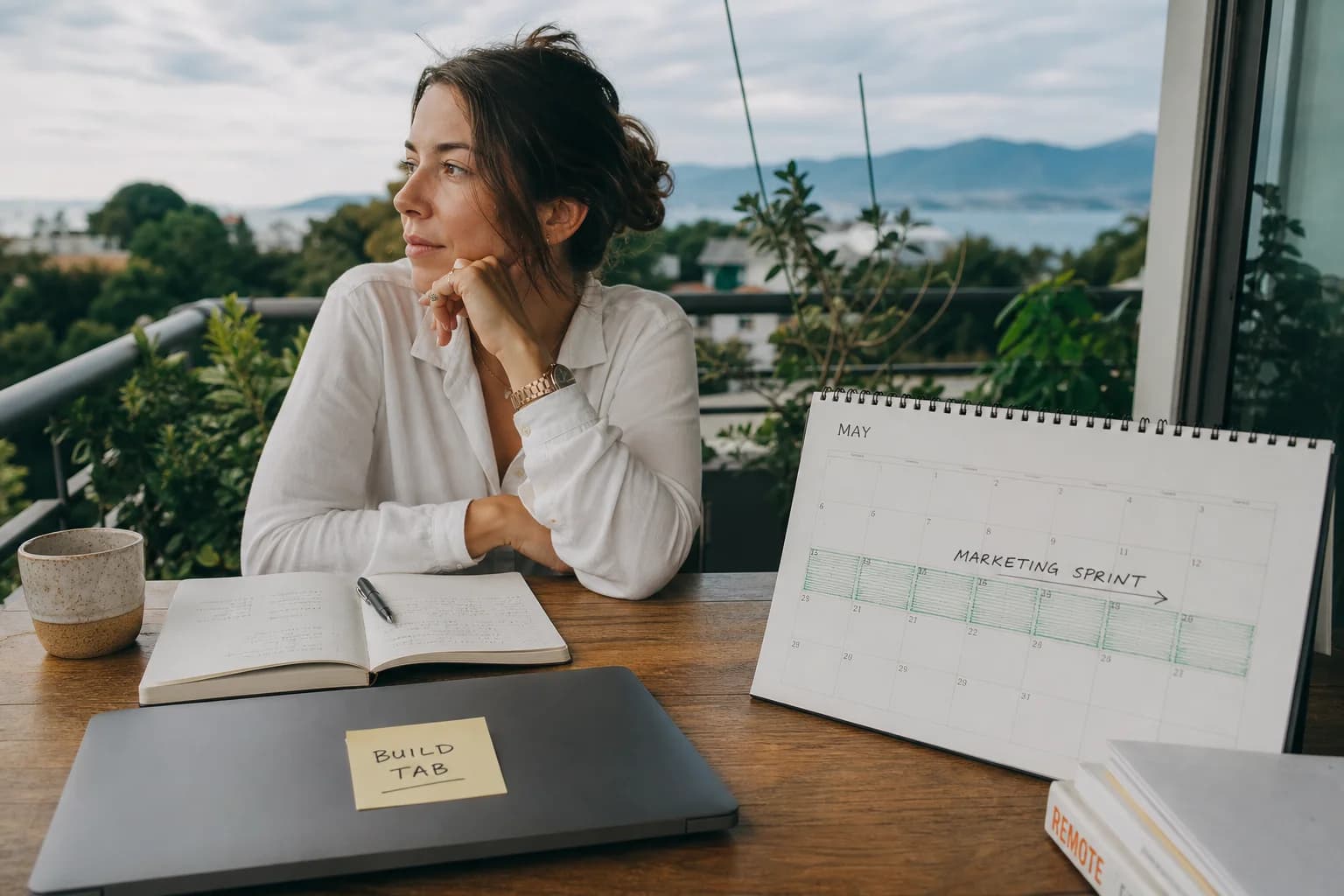Solo founder on a terrace desk with laptop and large monitor showing a kanban board beside a handwritten thirty-day sprint calendar and forest valley view